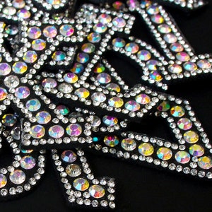 May include: Close-up of black fabric appliques densely covered with iridescent rhinestones. The rhinestones reflect a rainbow of colours. The appliques are arranged in a decorative pattern against a black background.