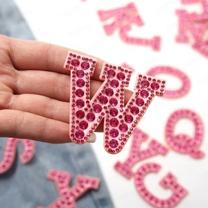 May include: A pink letter 'W' with a rhinestone design is held in a hand. The letter is surrounded by smaller pink rhinestones and larger pink rhinestones in the centre. Other pink rhinestone letters are in the background.