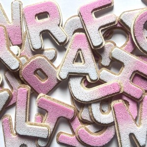 May include: A collection of individual alphabet letters featuring a textured, fuzzy surface with a gradient colour effect transitioning from white to pink. Each letter is outlined with a gold-coloured border, creating a distinct edge for every character.