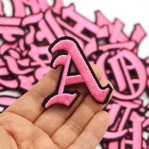 May include: An embroidered patch in the shape of the letter A, featuring bright pink thread with a thick black border. The letter is designed in a stylised gothic font with sharp, pointed edges and a textured surface from the stitching.