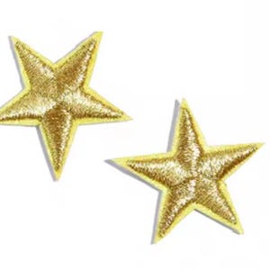 May include: Two gold star-shaped embroidered patches with yellow borders. The stars have a metallic sheen against a white background. These patches could be used for clothing or craft projects.