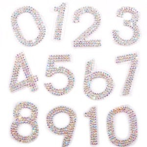 May include: A set of rhinestone-covered numbers from zero to zero. Each number is outlined with a clear border and is covered in iridescent rhinestones. The numbers are arranged on a white background, ideal for craft projects or embellishments.