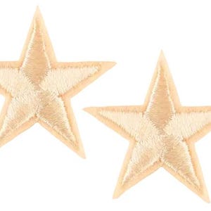 May include: Two embroidered star patches in a light tan colour. Each star has a five-point design with a detailed stitch pattern. The patches are likely for decorative purposes, such as on clothing or accessories.
