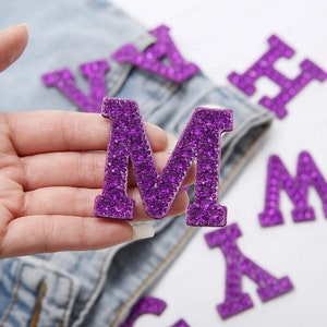 May include: A hand holding a purple, rhinestone-covered letter "M", with other letters visible. These decorative letters are likely for crafts or embellishments, featuring a sparkling, eye-catching design.