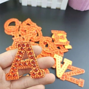 May include: An orange letter "A" covered in orange rhinestones, held in a hand, with other orange letters in the background. The letters are likely for crafts or personalisation. The letters are in the shape of the alphabet.