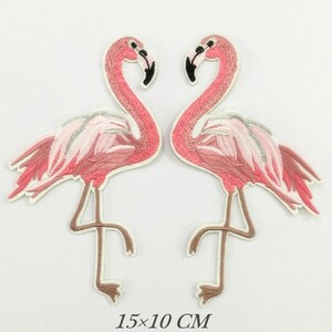 May include: Two embroidered flamingo patches. The flamingos are pink with white and silver accents on their wings. The patches are 15 cm by 10 cm.