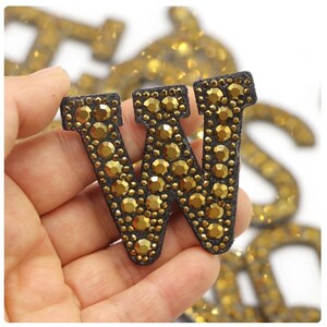 May include: A gold-coloured letter "W" patch, embellished with numerous small, round, gold-coloured rhinestones. The letter is outlined in black fabric. Other gold letters are visible in the background.