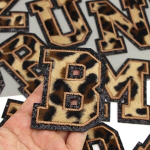 May include: A close-up of a letter "B" patch with a leopard print design. The patch has a black glitter border and a gold-coloured outline. Other letter patches are visible in the background.