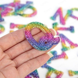 May include: A decorative letter "Q" in a rainbow gradient, adorned with iridescent beads and rhinestones. The letter is held in a hand, with other letters visible in the background. The letter's colours include yellow, green, blue, and purple.