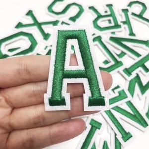 May include: Green embroidered letter "A" patch with a white border. The patch is held in a hand, with other letter patches in the background. The patches are likely for clothing or crafts.