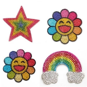 May include: Four rhinestone patches: a star, two smiling flower designs, and a rainbow with clouds. The star is red with yellow and green accents. The flowers have colourful petals and smiling faces. The rainbow is multi-coloured.