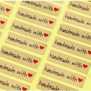 May include: A sheet of rectangular tan adhesive labels arranged in a grid pattern. Each label features the black printed text "handmade with" followed by a small red heart icon. The labels are presented on a light yellow backing sheet.