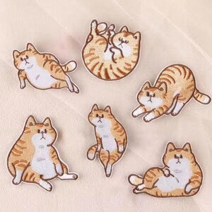May include: Six embroidered cat patches in different poses. The cats are orange and white with brown stripes. The patches show cats lying down, sitting, and stretching. Each patch is a different shape, suitable for clothing or accessories.