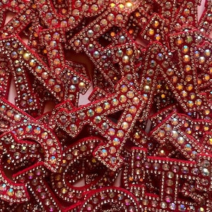 May include: A collection of red fabric letter patches densely covered in shimmering, iridescent rhinestones. Each letter features a bold, block-style font completely encrusted with sparkling crystals that reflect light, creating a textured, multi-dimensional appearance across the overlapping pieces.