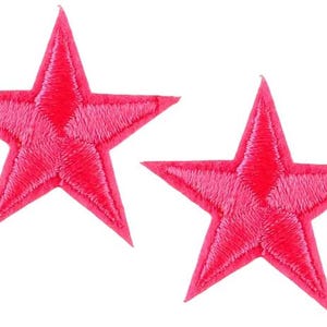 May include: Two embroidered pink star patches. Each star has five points and is filled with textured stitching. The stars are a vibrant shade of pink against a white background.