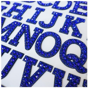May include: Close-up of dazzling blue rhinestone letters on a white surface. The letters, in a bold serif font, are covered in small, reflective rhinestones. The arrangement suggests alphabet tiles or decorative elements, ideal for crafts.