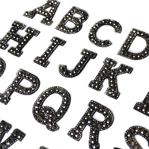 May include: A selection of alphabet letters, each embellished with small, dark grey rhinestones. The letters are in a sans-serif style and are displayed against a white backdrop. The letters include A, B, C, D, H, I, J, K, P, Q, and R.