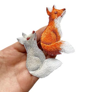 May include: Two embroidered fox patches. One is orange and white, the other is white. Both are looking upwards with detailed stitching. These patches could be used to decorate clothing or accessories.