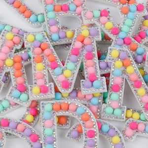 May include: A selection of decorative letters, each adorned with pastel-coloured beads and sparkling rhinestones. The letters feature a variety of colours, such as pink, orange, yellow, green, and blue. The letters are suitable for crafts.