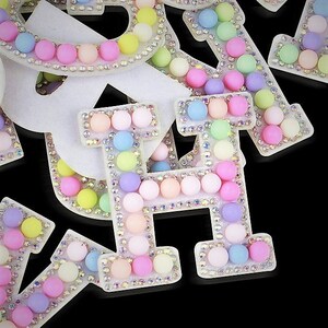 May include: Close-up of pastel-coloured letter embellishments. Each letter is decorated with small, round, pastel-coloured beads and surrounded by iridescent rhinestones. The letters are white with a black background.