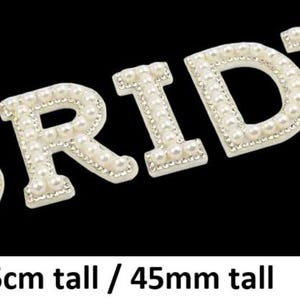 May include: Ivory-coloured 'BRIDE' letters, each decorated with small, round pearls, are set against a black background. The letters are 4.5cm tall. Ideal for wedding decorations.