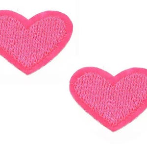 May include: Two embroidered pink heart patches. Each heart has a textured fill and a darker pink border. The patches are set against a white background.
