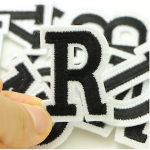 May include: Black embroidered letter "R" patch with a white border. The patch is part of a set of letters and numbers, ideal for customising clothing or accessories. The patches are made of fabric and have an iron-on backing.