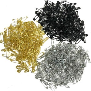 May include: A selection of safety pins in gold, black, and silver. The pins are arranged in separate piles against a white backdrop. The pins are made of metal and feature a traditional safety pin design.