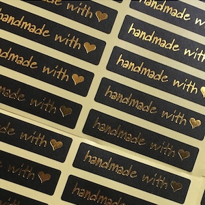 May include: Close-up of black rectangular stickers with gold text that reads "handmade with" and a gold heart. The stickers are arranged in rows and columns on a light-coloured surface.