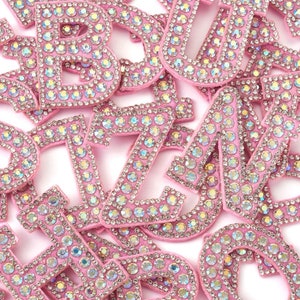 May include: A selection of pink fabric letters, each adorned with shimmering rhinestones. The letters are arranged on a white background, highlighting the sparkling details and the range of the alphabet. Suitable for crafting and customisation.