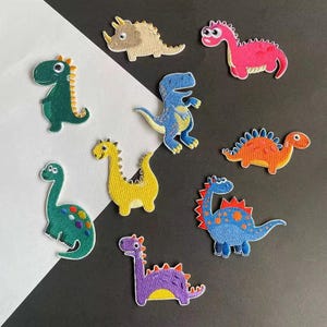 May include: A selection of embroidered dinosaur patches in assorted colours and designs. The patches feature detailed stitching and a range of hues, including green, blue, yellow, pink, and purple. They are displayed against a black and white backdrop.