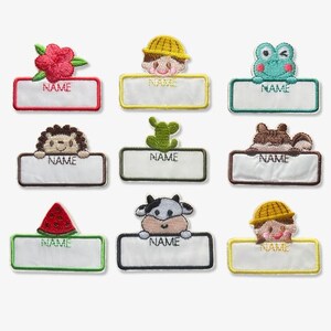 May include: Nine embroidered name tags featuring animal and floral designs. Each tag has a white rectangular space for a name, framed by different coloured borders. Designs include a red flower, a boy in a yellow hat, a frog, a hedgehog, a cactus, a bear, a watermelon, a cow, and a girl in a yellow hat.
