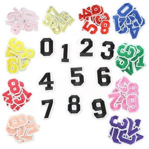 May include: A collection of number patches in a variety of colours, including pink, gold, red, blue, yellow, green, and purple. Each number has a black outline and a white border. Numbers 0 to 9 are shown.