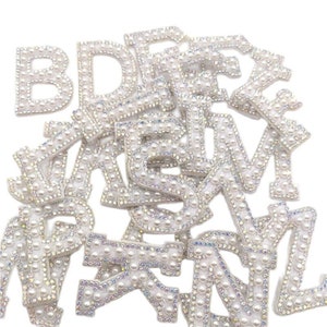 May include: A selection of white alphabet letters, each embellished with pearls. The letters are outlined in white and filled with small, iridescent pearls. The letters are scattered on a white background, suitable for crafting or adding a decorative touch.