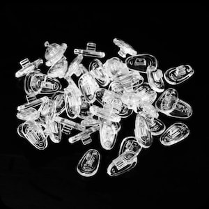 May include: A pile of clear, transparent eyeglass nose pads. The pads are made of a smooth, glossy material and are designed to fit various eyeglass frames. They are scattered on a black surface.