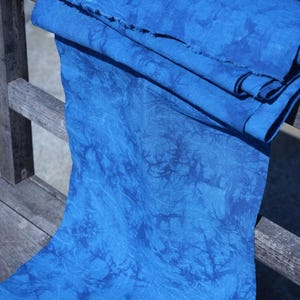May include: A bolt of vibrant blue fabric with a textured, tie-dye-like pattern. The fabric is draped over a wooden surface, showcasing its rich color and unique design. The edges of the fabric are visible, highlighting its texture.