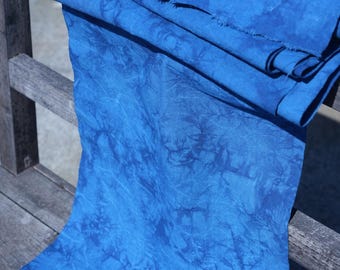 May include: A bolt of vibrant blue fabric with a textured, tie-dye-like pattern. The fabric is draped over a wooden surface, showcasing its rich color and unique design. The edges of the fabric are visible, highlighting its texture.
