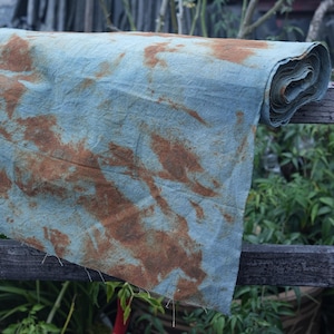 Puede incluir: Una tela azul con patrones de tinte de color óxido. La tela está doblada y enrollada, y está sentada en una valla de madera.