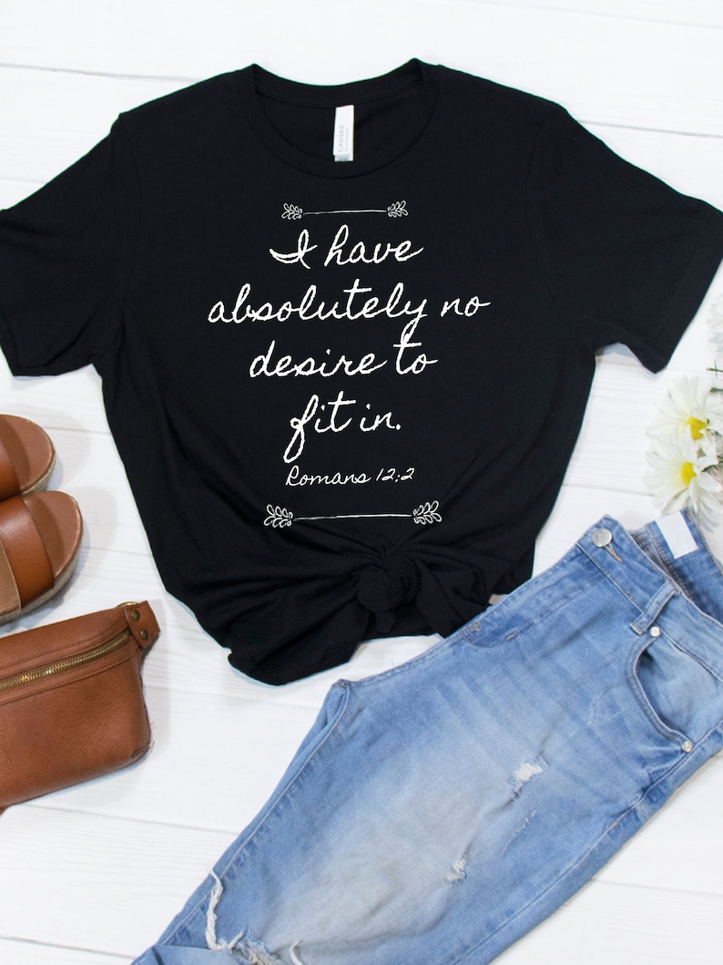 May include: Black t-shirt with white text that reads "I have absolutely no desire to fit in. Romans 12:2". The shirt is styled with a knot in the front and is paired with blue jeans and a brown leather fanny pack.