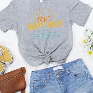 May include: A gray t-shirt with a graphic that says "Don't Throw Shade" with a sun and waves underneath. The text "Ephesians 4:29" is also included on the shirt. The shirt is tied in a knot and is laying on a white wooden surface with a brown leather fanny pack, blue jeans, and yellow sneakers.