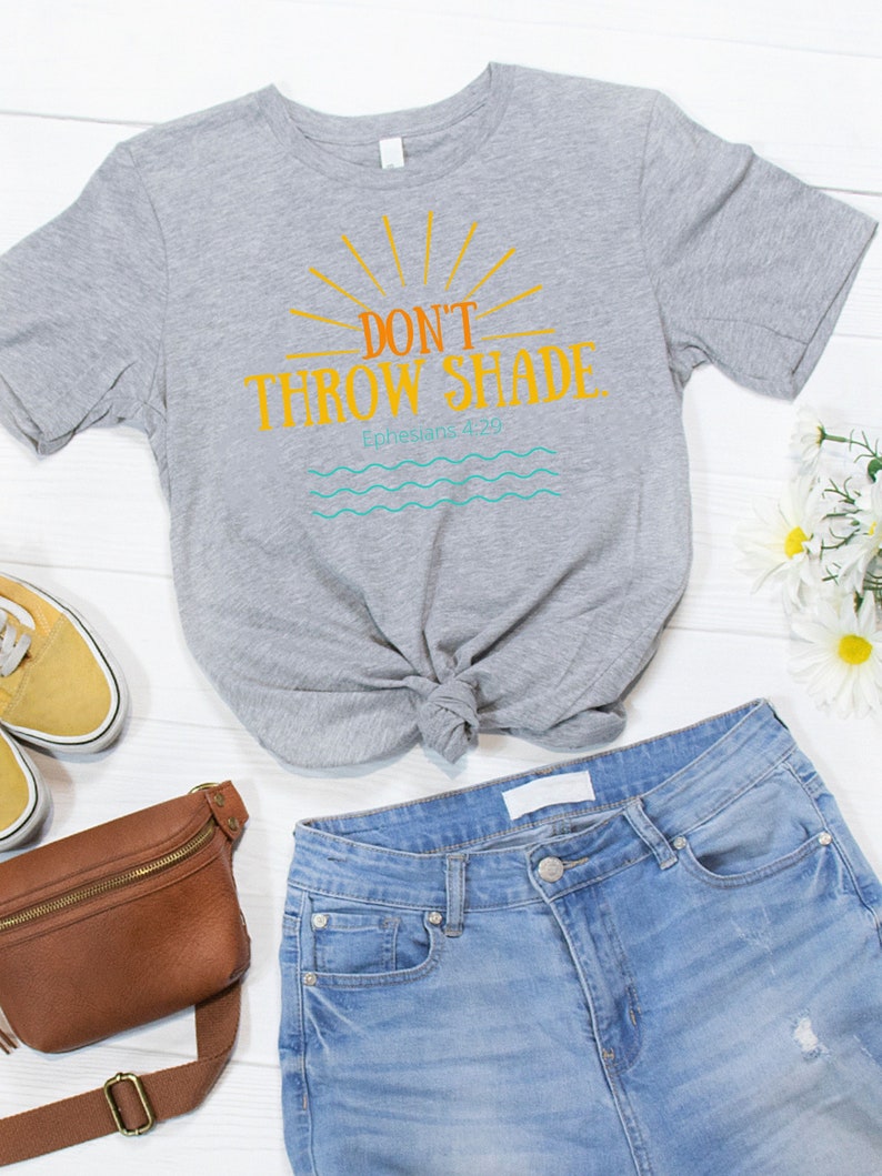 May include: A gray t-shirt with a graphic that says "Don't Throw Shade" with a sun and waves underneath. The text "Ephesians 4:29" is also included on the shirt. The shirt is tied in a knot and is laying on a white wooden surface with a brown leather fanny pack, blue jeans, and yellow sneakers.