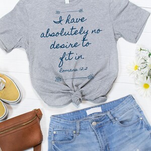 May include: Grey t-shirt with the text "I have absolutely no desire to fit in. Romans 12:2" printed on it. The shirt is tied in a knot and is lying on a white wooden surface with a brown leather fanny pack, a pair of yellow trainers, and white flowers.