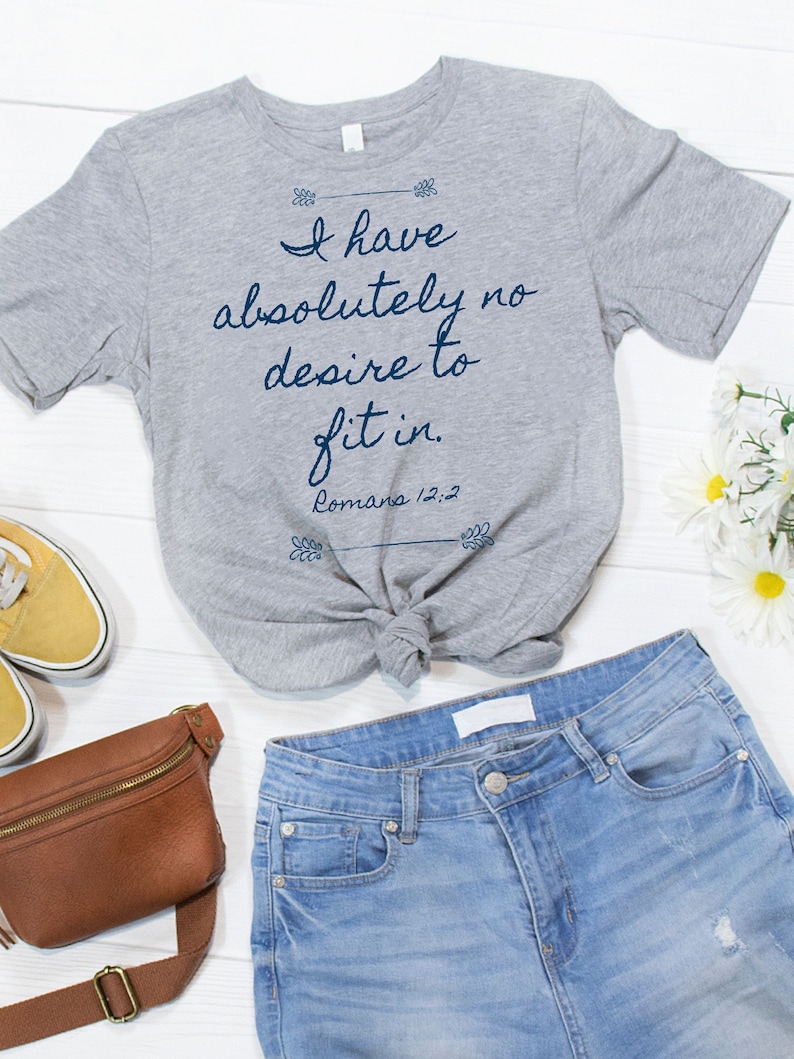 May include: Grey t-shirt with the text "I have absolutely no desire to fit in. Romans 12:2" printed on it. The shirt is tied in a knot and is lying on a white wooden surface with a brown leather fanny pack, a pair of yellow trainers, and white flowers.