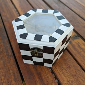 May include: A hexagonal wooden box with a checkered black and white pattern. The box has a clear glass lid and a brass latch.