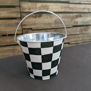 May include: A small metal bucket with a black and white checkered pattern. The bucket has a silver handle and is sitting on a dark gray surface.