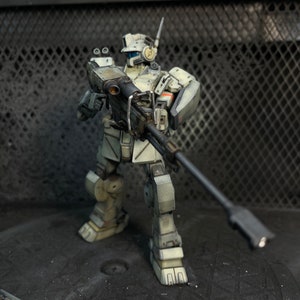 May include: A white and gray robot action figure holding a large black gun. The robot is standing on a gray surface with a black metal grate in the background.
