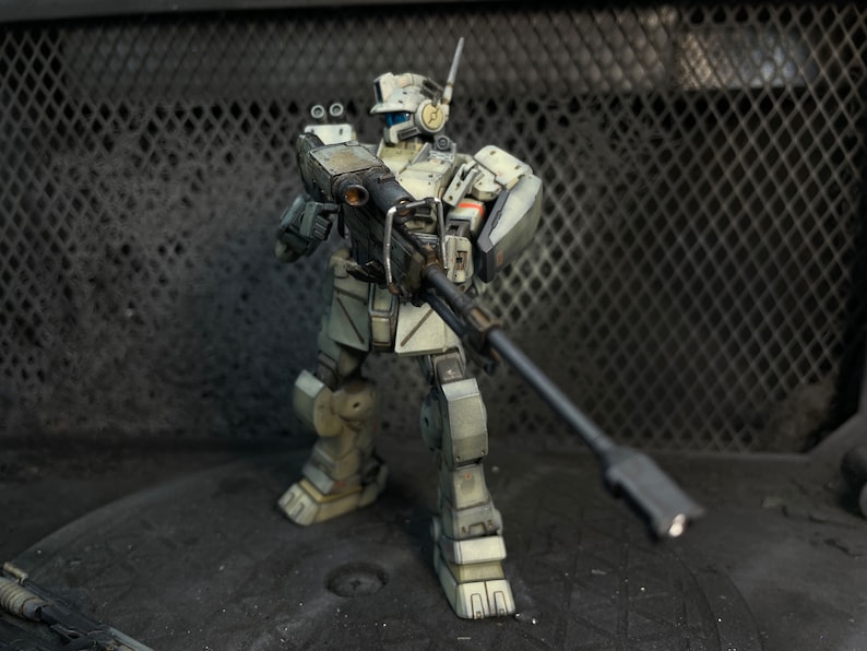 May include: A white and gray robot action figure holding a large black gun. The robot is standing on a gray surface with a black metal grate in the background.