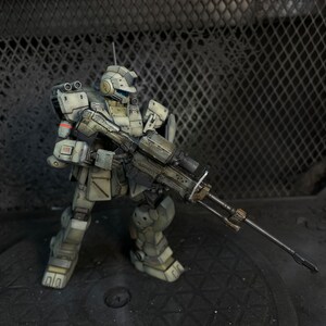 May include: A white and gray robot action figure holding a large black rifle. The robot is standing on a black surface with a metal grate in the background.