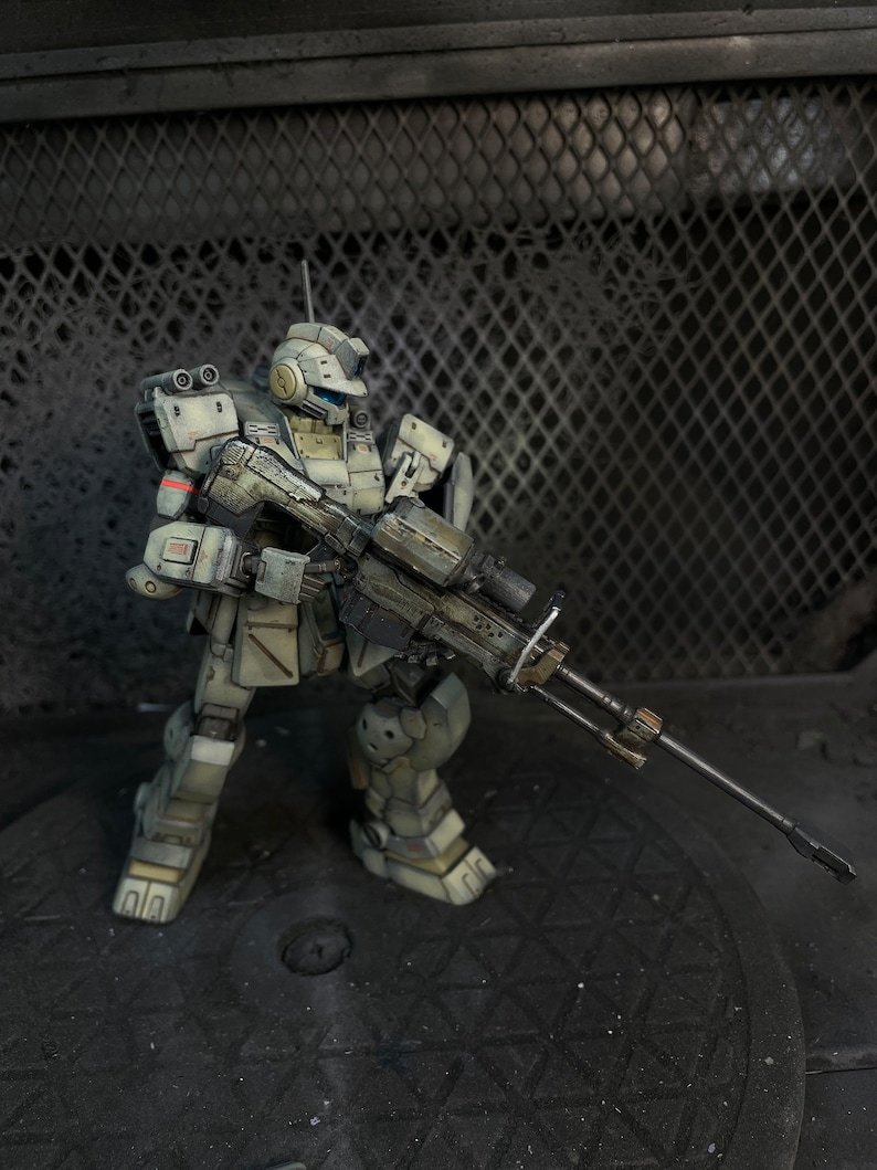 May include: A white and gray robot action figure holding a large black rifle. The robot is standing on a black surface with a metal grate in the background.