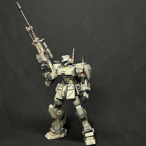 May include: A white and gray robot action figure holding a large rifle. The robot has a helmet with a visor and a detailed design. The figure is posed in a standing position with the rifle raised.